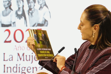 La presidenta Claudia Sheinbaum sostiene y muestra su libro Diario de una transición histórica durante su presentación en Palacio Nacional. Al fondo se observa un cartel con la leyenda “2025, Año de la Mujer Indígena”.