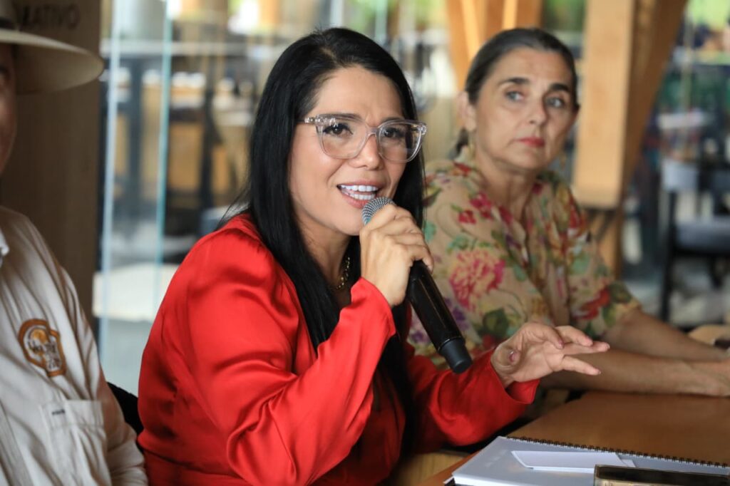 Katia Ornelas, de blusa roja toma el micrófono en conferencia junto a otros asistentes.