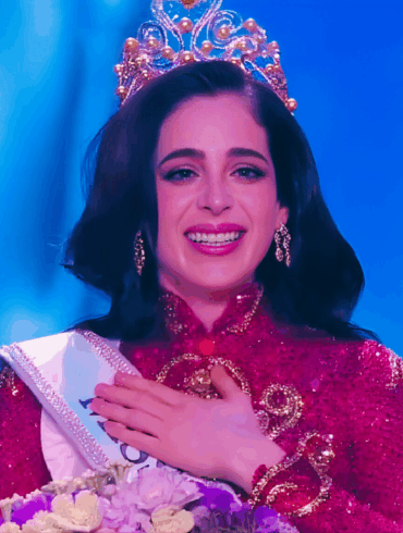 Mujer coronada con traje rojo y corona dorada, sonriendo emocionada en el escenario de Miss Universe 2025.