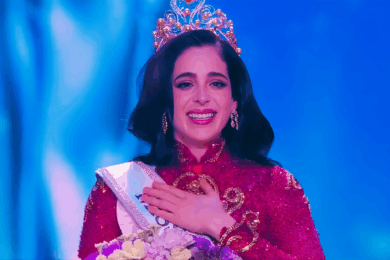 Mujer coronada con traje rojo y corona dorada, sonriendo emocionada en el escenario de Miss Universe 2025.