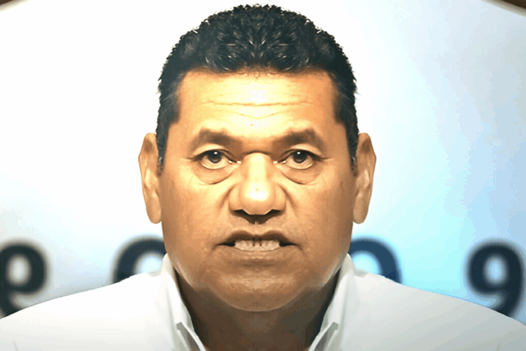 Primer plano de Javier May con camisa blanca, mirando de frente, mientras ofrece un mensaje en un evento público, con fondo desenfocado.