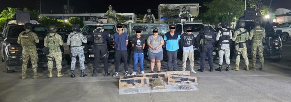 Elementos de fuerzas federales y estatales rodean a cinco personas detenidas, colocadas frente a varias patrullas durante un operativo nocturno; en el suelo se observan armas largas aseguradas, una subametralladora y paquetes con presunta droga.