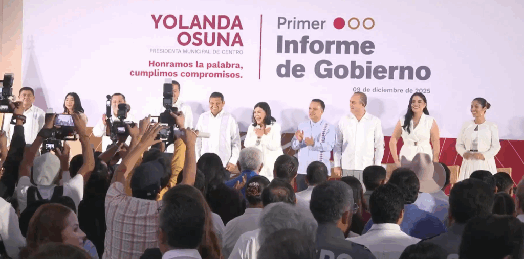 Yolanda Osuna y su equipo en el escenario durante el Primer Informe de Gobierno, acompañados por funcionarios y ante un público que documenta el momento con teléfonos y cámaras.