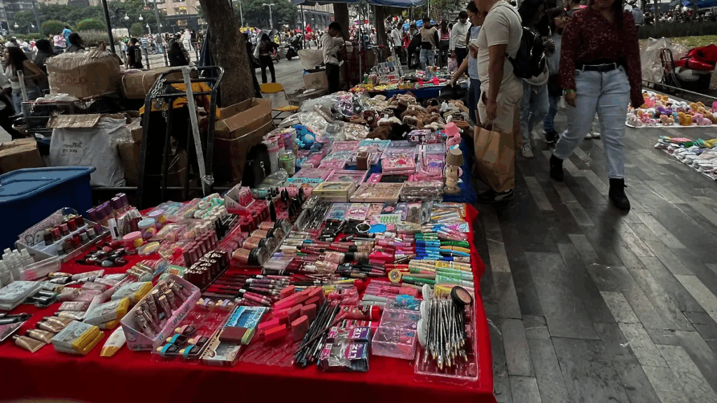 Puesto ambulante con cosméticos, bisutería y artículos variados instalado sobre una banqueta transitada, donde peatones caminan entre mercancía y cajas apiladas en la vía pública.
