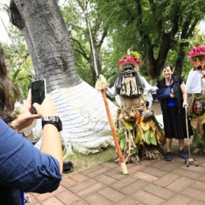 “Una mujer fotografía a dos participantes disfrazados con trajes tradicionales del Carnaval de Tenosique que posan junto a una visitante en un parque rodeado de árboles.”