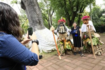 “Una mujer fotografía a dos participantes disfrazados con trajes tradicionales del Carnaval de Tenosique que posan junto a una visitante en un parque rodeado de árboles.”