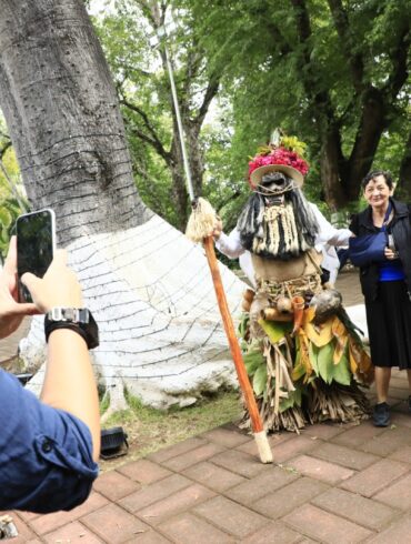 “Una mujer fotografía a dos participantes disfrazados con trajes tradicionales del Carnaval de Tenosique que posan junto a una visitante en un parque rodeado de árboles.”