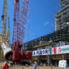 Estructura industrial de Pemex en construcción con grúas de gran tonelaje, tubería de coque en maniobra de instalación y trabajadores supervisando la obra en una planta petrolera.