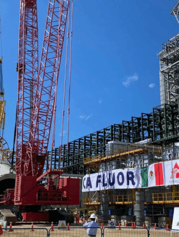 Estructura industrial de Pemex en construcción con grúas de gran tonelaje, tubería de coque en maniobra de instalación y trabajadores supervisando la obra en una planta petrolera.