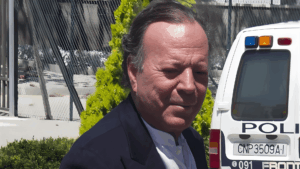 “Primer plano de un hombre de cabello canoso caminando al aire libre junto a una patrulla policial; detrás aparecen rejas metálicas y vegetación.”