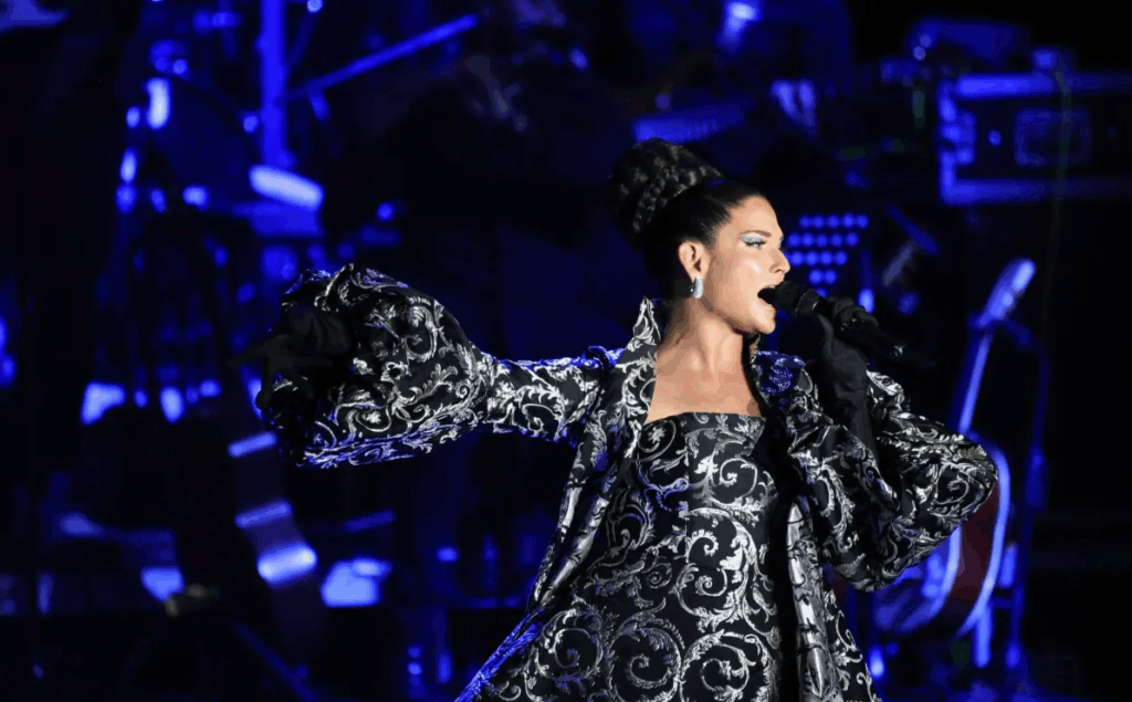 Natalia Jiménez canta en un concierto en vivo, iluminada en tonos azules, con un vestido bordado de estilo ranchero-pop.
