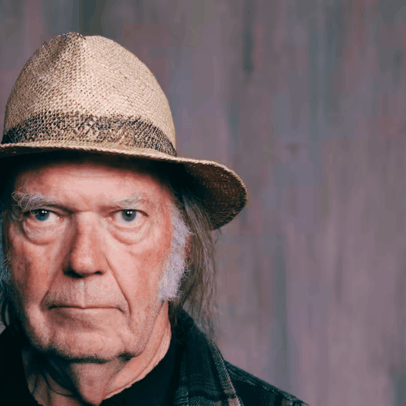 Retrato cerrado de Neil Young, con sombrero de paja y gesto serio, mirando a cámara en fondo gris.