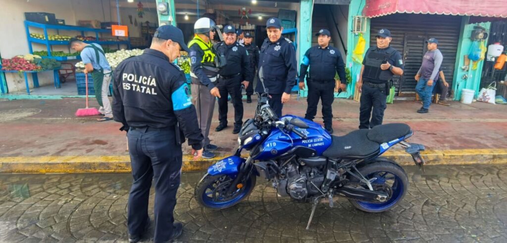 Grupo de elementos de la Policía Estatal y Policía Estatal de Caminos inspecciona una motocicleta oficial durante un operativo en zona comercial del mercado, con locatarios al fondo.