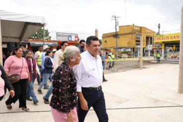 El gobernador Javier May Rodríguez camina junto a una vecina durante la supervisión de obras de equipamiento urbano en Cunduacán, mientras personal técnico y habitantes acompañan el recorrido en zona intervenida.