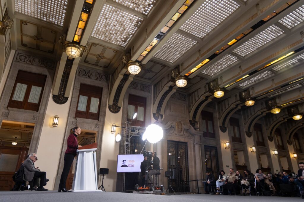 Funcionaria ofrece un mensaje desde el podio en un salón histórico con arquitectura iluminada, rodeada de prensa y equipo técnico.