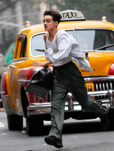Timothée Chalamet corre despreocupado por una calle de Nueva York en una escena de Marty Supreme, caracterizado como Marty Mauser, con camisa abierta, lentes y un taxi amarillo detrás de él.