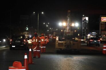 Trabajadores municipales con chalecos reflejantes realizan bacheo nocturno en el periférico de Villahermosa con conos de señalización y tráfico vehicular circulando a un costado.