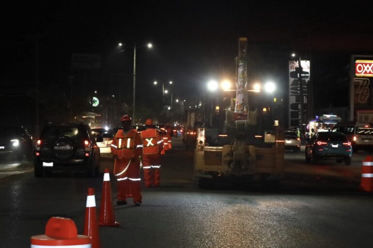 Trabajadores municipales con chalecos reflejantes realizan bacheo nocturno en el periférico de Villahermosa con conos de señalización y tráfico vehicular circulando a un costado.