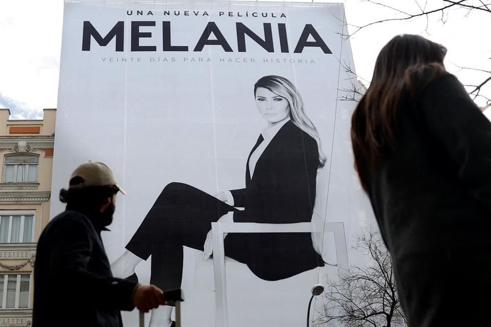 Cartel monumental de la película documental Melania con retrato en blanco y negro de Melania Trump instalado en una avenida, mientras peatones caminan frente al anuncio.