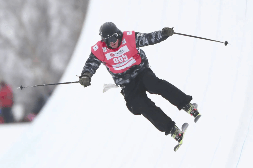 Esquiador en competencia, suspendido en el aire durante un salto, con peto rojo y bastones extendidos sobre un fondo blanco de nieve.