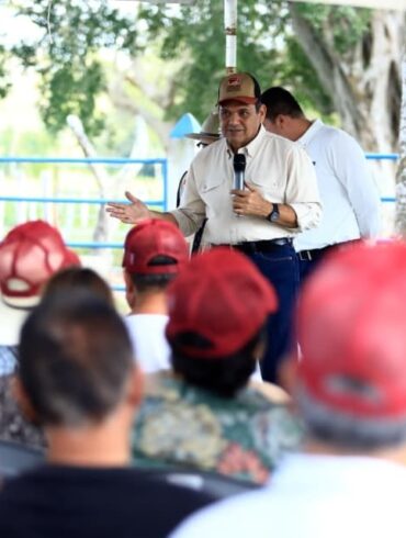Fotografía del gobernador de Tabasco, Javier May Rodríguez, vistiendo camisa clara y gorra café, hablando por micrófono frente a un grupo de ganaderos que escuchan atentos.