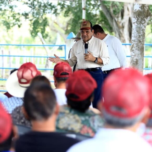 Fotografía del gobernador de Tabasco, Javier May Rodríguez, vistiendo camisa clara y gorra café, hablando por micrófono frente a un grupo de ganaderos que escuchan atentos.