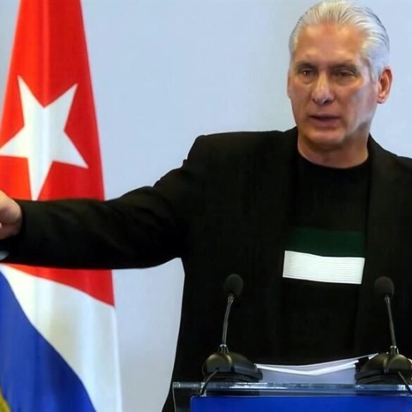 Miguel Díaz-Canel habla en conferencia de prensa frente a la bandera cubana mientras señala con el brazo extendido.