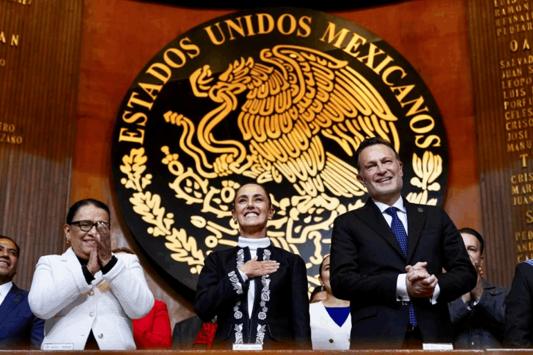 Claudia Sheinbaum junto a integrantes del gabinete y autoridades federales aplauden frente al escudo nacional durante el acto oficial.