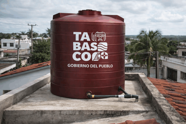 Tinaco color vino de 450 litros con logotipo del Gobierno de Tabasco instalado en la azotea de una vivienda, con tubería conectada y vista urbana al fondo en Villahermosa.