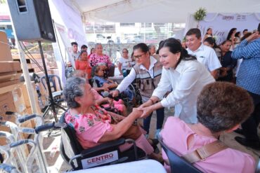 Presidenta municipal Yolanda Osuna Huerta saluda a adulta mayor en silla de ruedas durante la octava jornada de Centro +Cerca en la colonia Primero de Mayo, Villahermosa, Tabasco. Al fondo, módulos de entrega de aparatos funcionales y asistentes al evento.
