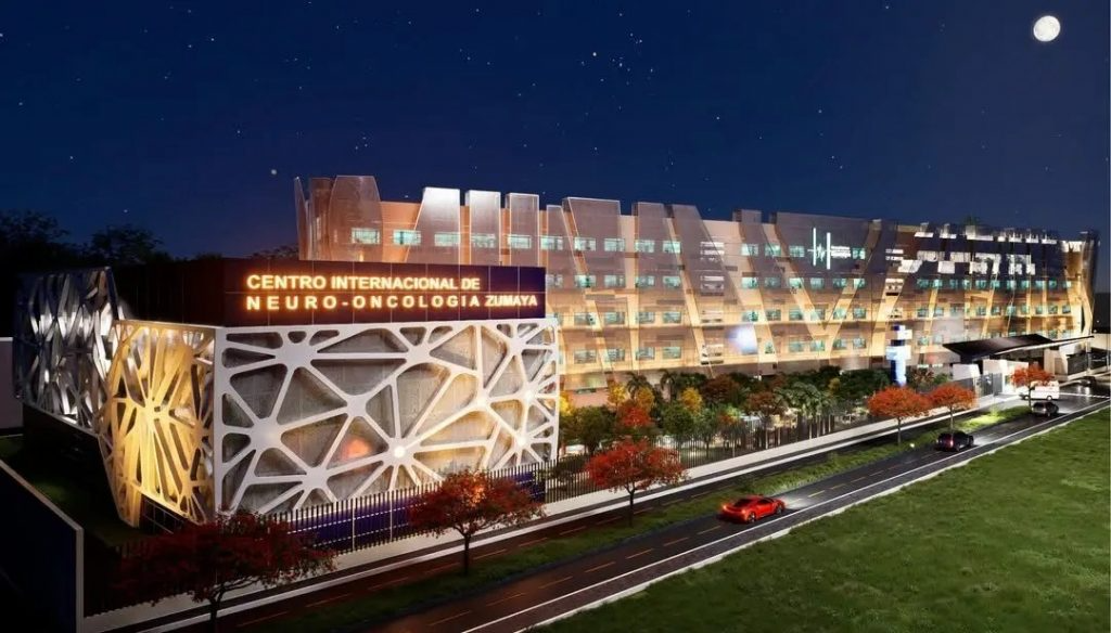 Render nocturno del Centro Internacional de Neuro-Oncología Zumaya en Villahermosa, con fachada paramétrica iluminada en ámbar y acceso vehicular al frente