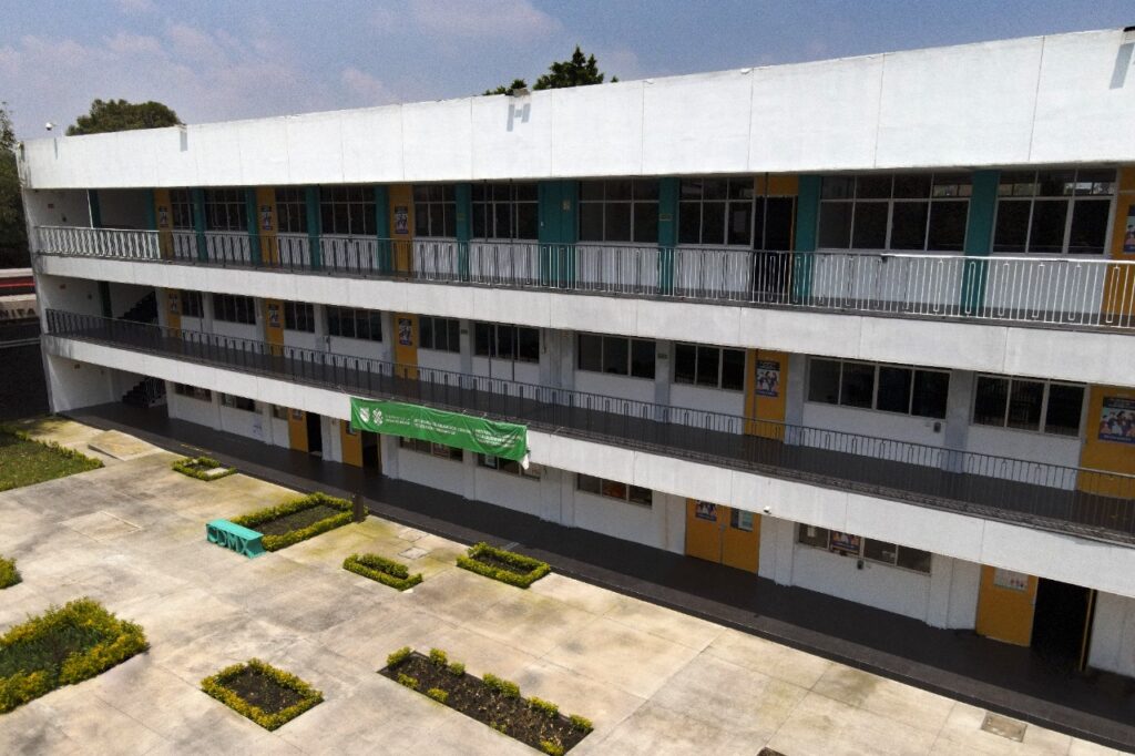 Vista aérea del patio interior de una sede de la Universidad Nacional Rosario Castellanos en la Ciudad de México, con banner institucional verde y áreas jardinadas