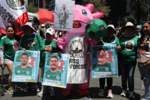 Persona disfrazada de axolotl con la leyenda México Crisis Forense, rodeada de carteles estilo carta FIFA con fotos de personas desaparecidas en Ecatepec