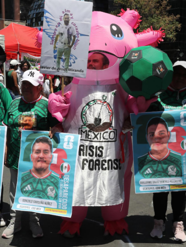 Persona disfrazada de axolotl con la leyenda México Crisis Forense, rodeada de carteles estilo carta FIFA con fotos de personas desaparecidas en Ecatepec