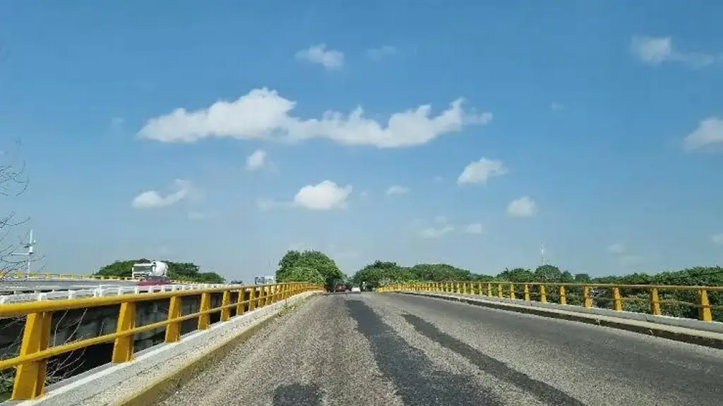 Puente carretero de dos carriles con barandal amarillo y vegetación a los costados, bajo cielo despejado con nubes dispersas
