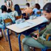 Estudiante consulta un teléfono celular durante una clase mientras sus compañeros atienden a la maestra
