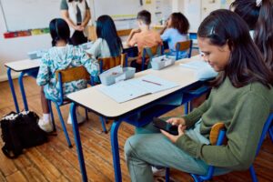 Estudiante consulta un teléfono celular durante una clase mientras sus compañeros atienden a la maestra