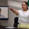 María Eliza Hernández rinde protesta mientras Alfonso Baca aparece a distancia en una pantalla.