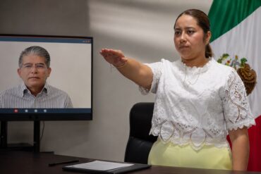 María Eliza Hernández rinde protesta mientras Alfonso Baca aparece a distancia en una pantalla.