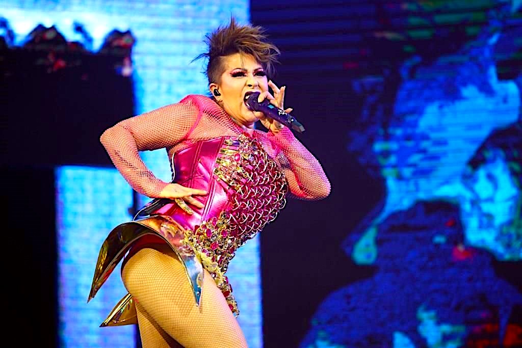 Alejandra Guzmán canta al micrófono durante un concierto, con vestuario rosa y dorado sobre escenario iluminado en azul