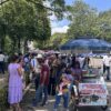 Multitud caminando bajo los árboles hacia el Estadio Olímpico de Villahermosa, junto a un carrito de pozol con sombrilla azul.