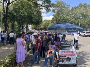 Multitud caminando bajo los árboles hacia el Estadio Olímpico de Villahermosa, junto a un carrito de pozol con sombrilla azul.