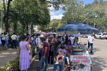 Multitud caminando bajo los árboles hacia el Estadio Olímpico de Villahermosa, junto a un carrito de pozol con sombrilla azul.