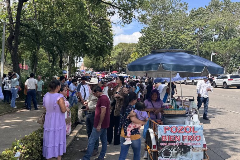 Multitud caminando bajo los árboles hacia el Estadio Olímpico de Villahermosa, junto a un carrito de pozol con sombrilla azul.