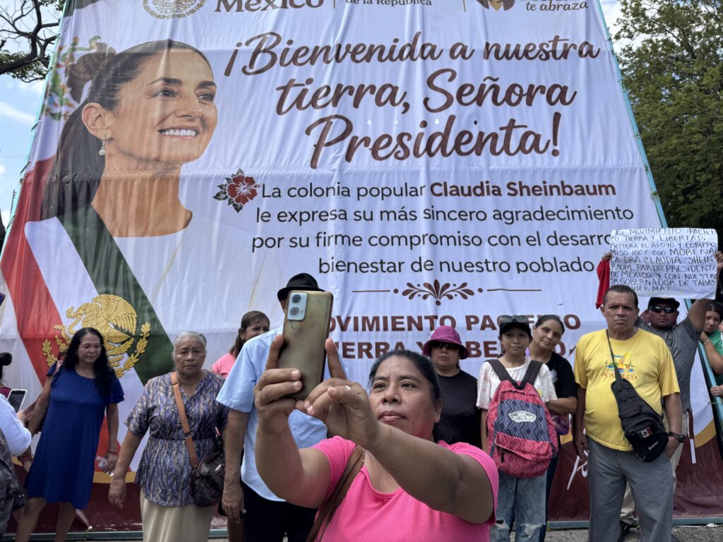 Mujer tomando una foto con su celular frente a una lona gigante de bienvenida a la presidenta Claudia Sheinbaum.