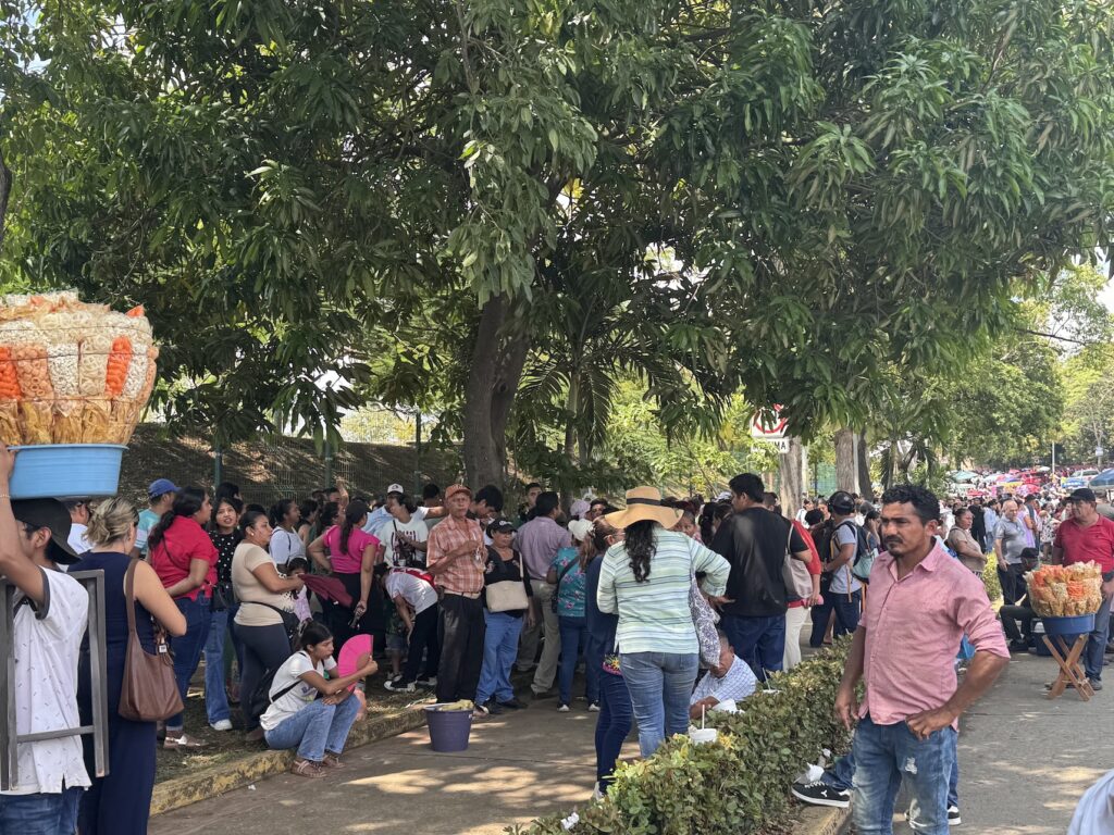 Asistentes resguardados del sol bajo árboles de mango afuera del estadio, con un vendedor de frituras a un lado.