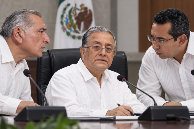 Composición visual de tres funcionarios y actores políticos en una mesa de sesión, con el secretario Ejecutivo del órgano electoral al centro bajo presión política.