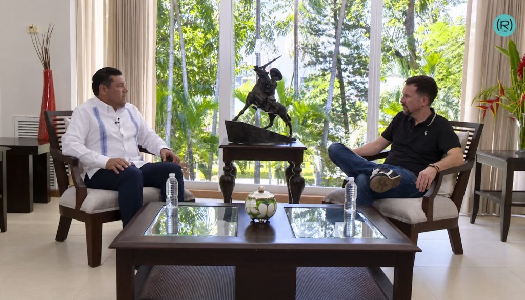Javier May conversa con un entrevistador mientras ambos permanecen sentados frente a frente.