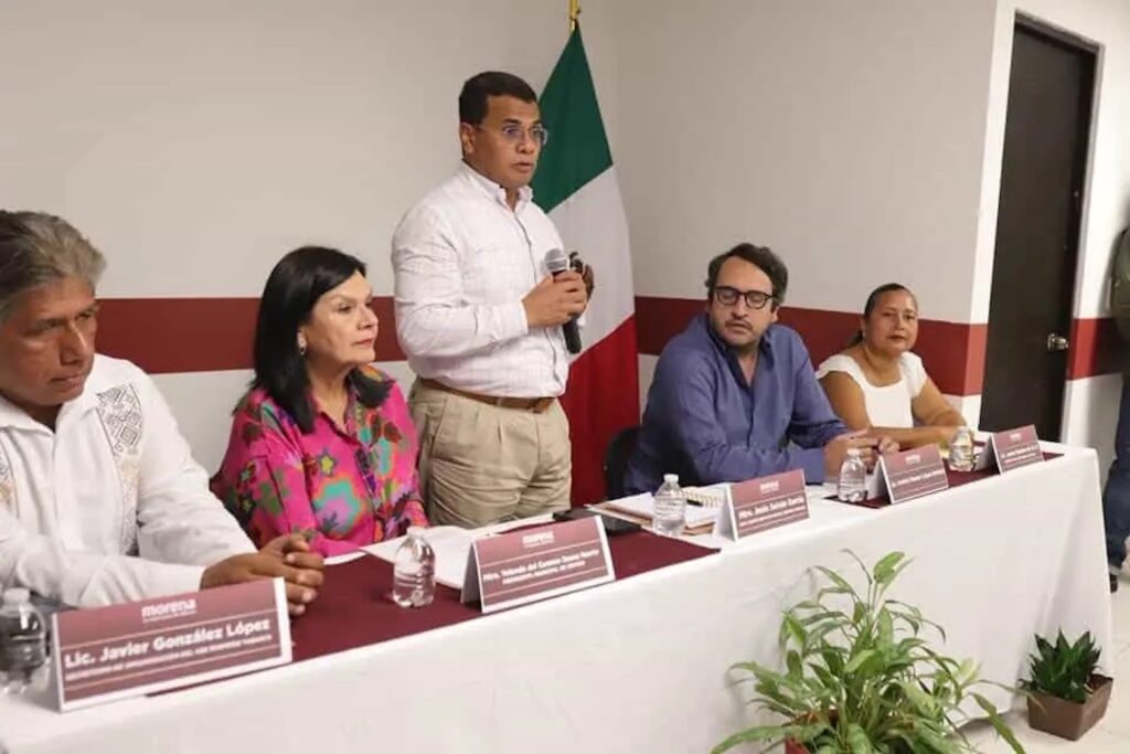 Jesús Selván García habla de pie con micrófono en una reunión de Morena en Tabasco, junto a la bandera de México