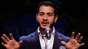 Abdul El-Sayed pronuncia un discurso ante un micrófono con ambas manos extendidas durante un acto público.